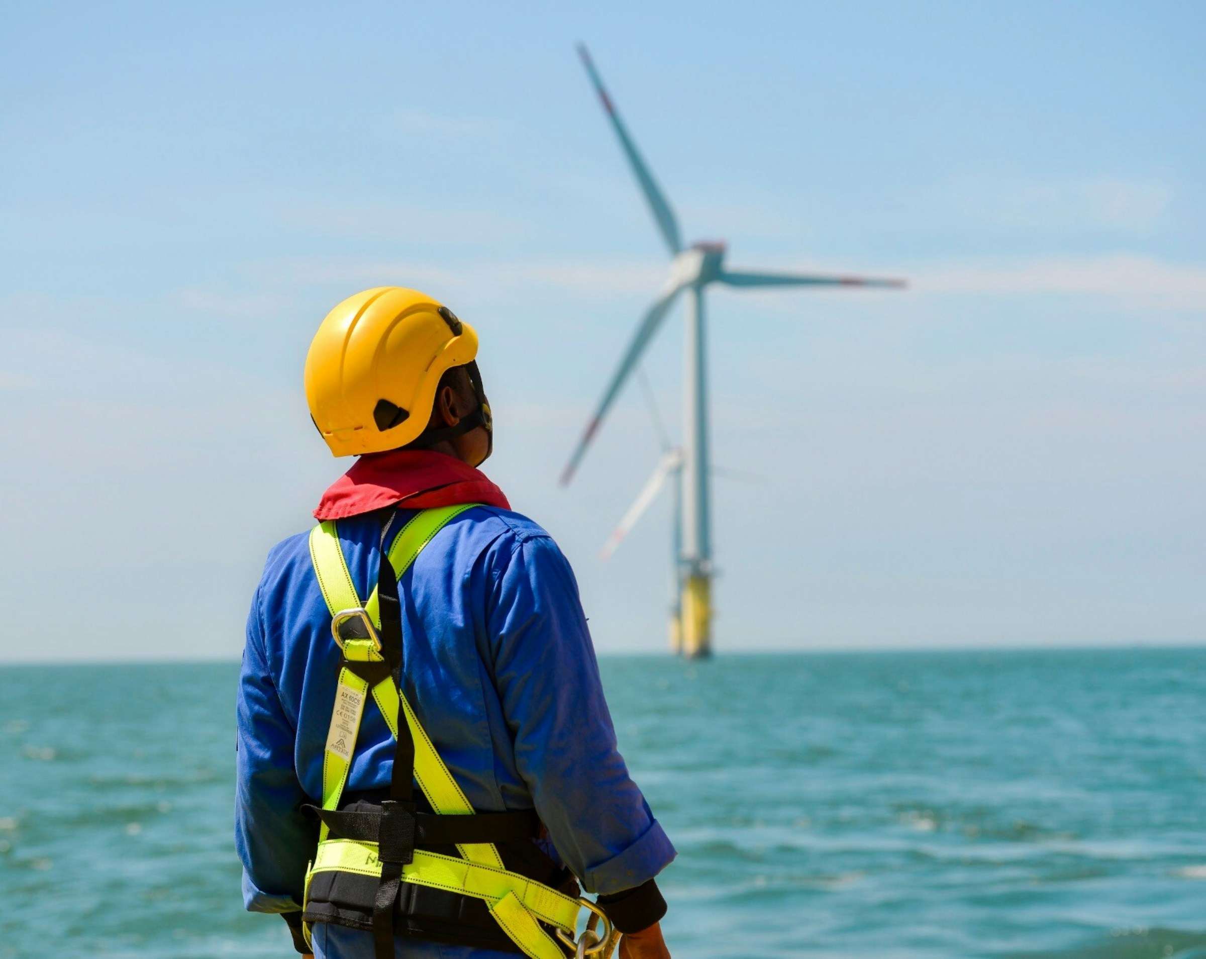 A field technician in a hard hat and safety harness watching over an offshore wind turbine
