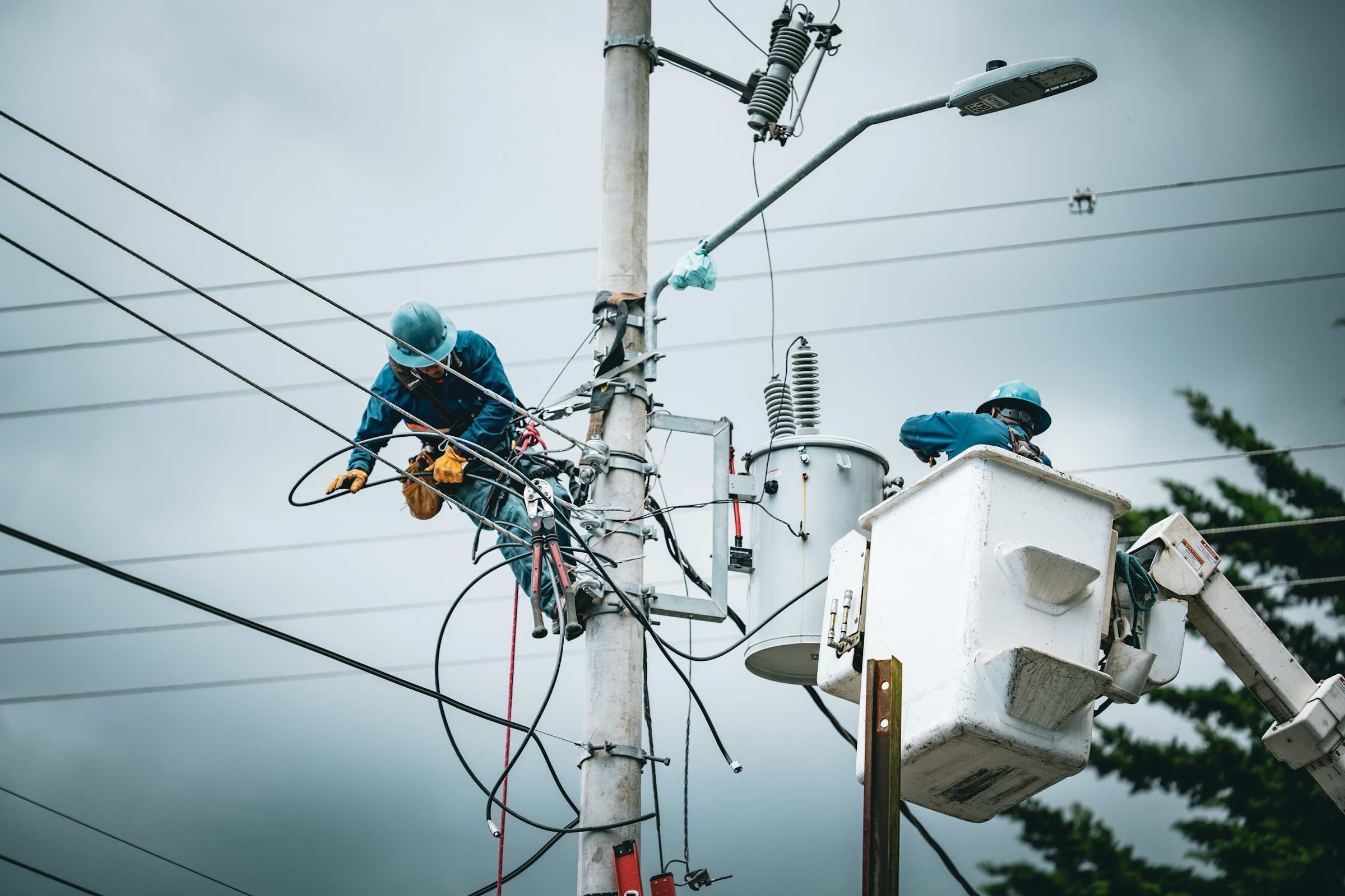 Two field technicians in hard hats working on a pole-top transformer, one on the pole and one in a bucket lift